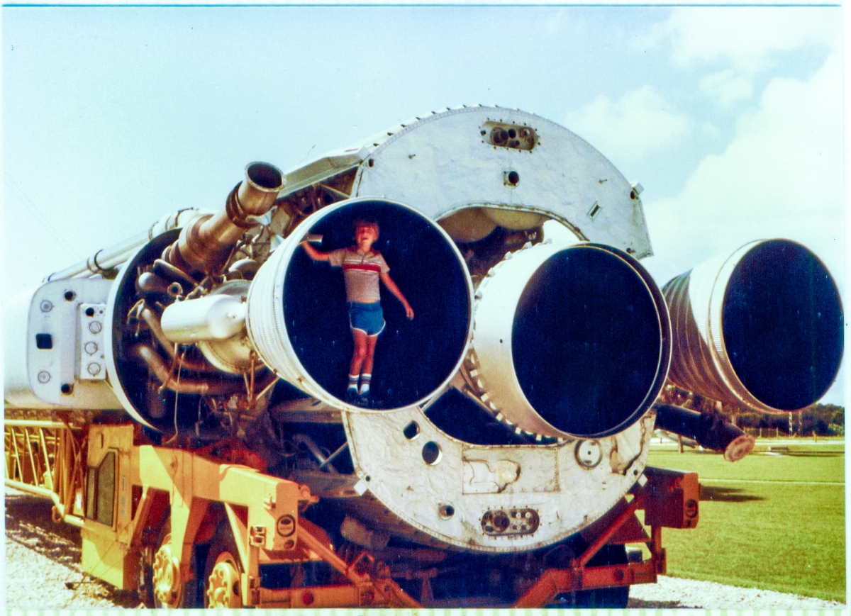 Kai MacLaren stands in one of the booster-engine nozzles of an Atlas ICBM (this is flight hardware and is not a replica) on display at Complex-26, Air Force Space Museum, Cape Canaveral, Florida. To the left of him in this frame, some of the metal covering which ordinarily encases the engine compartment of the Atlas has been removed, allowing a glimpse of the complexity of engine itself. The Atlas is resting on the “stretcher” by which it was transported to the pad, where it would then be lifted into a vertical position above the flame bucket, and prepared for launch.